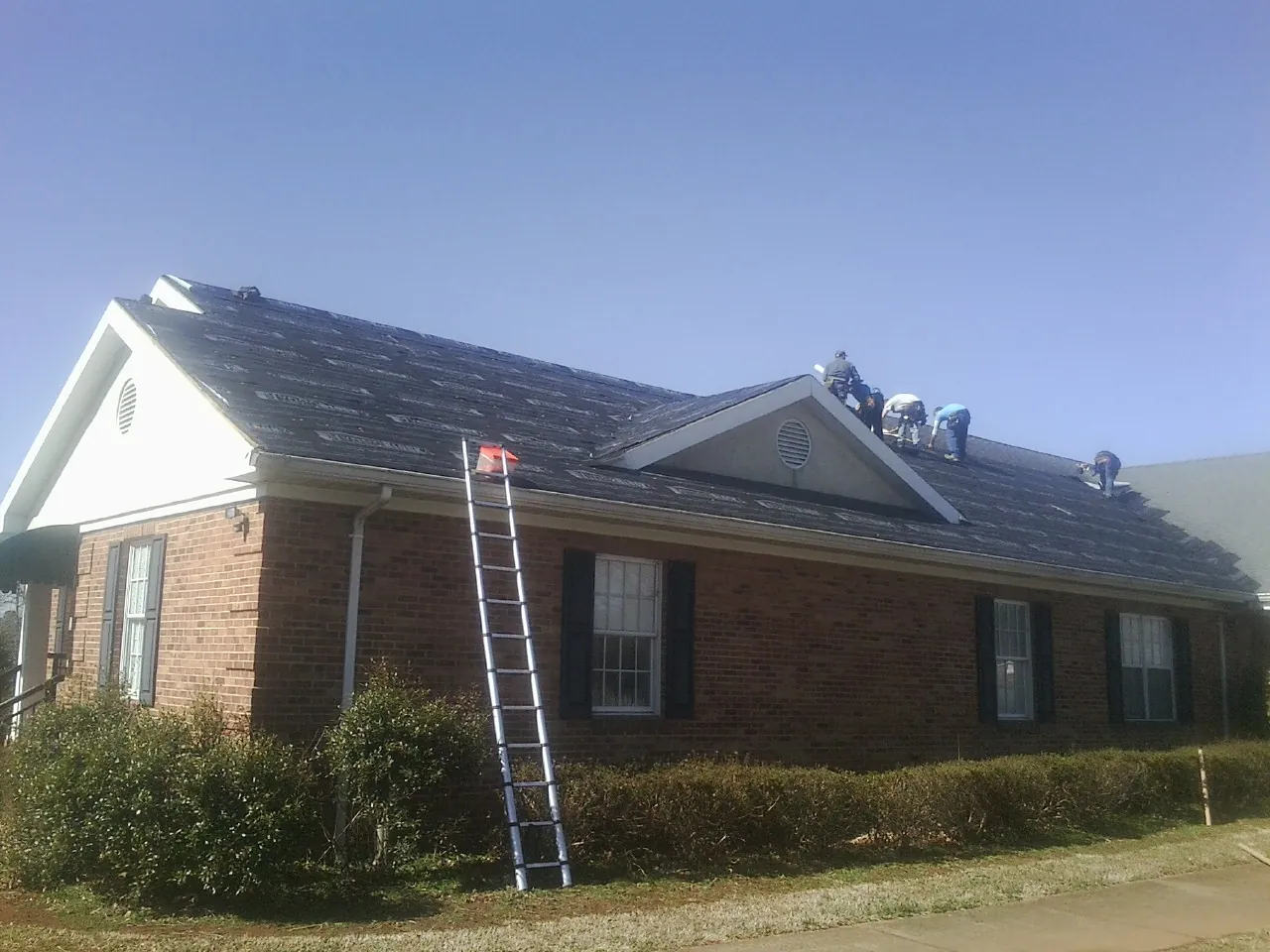 Crew tearing shingles off a brick ranch roof with a ladder set — Metal Roof Repair work by our crew in Sugarcreek