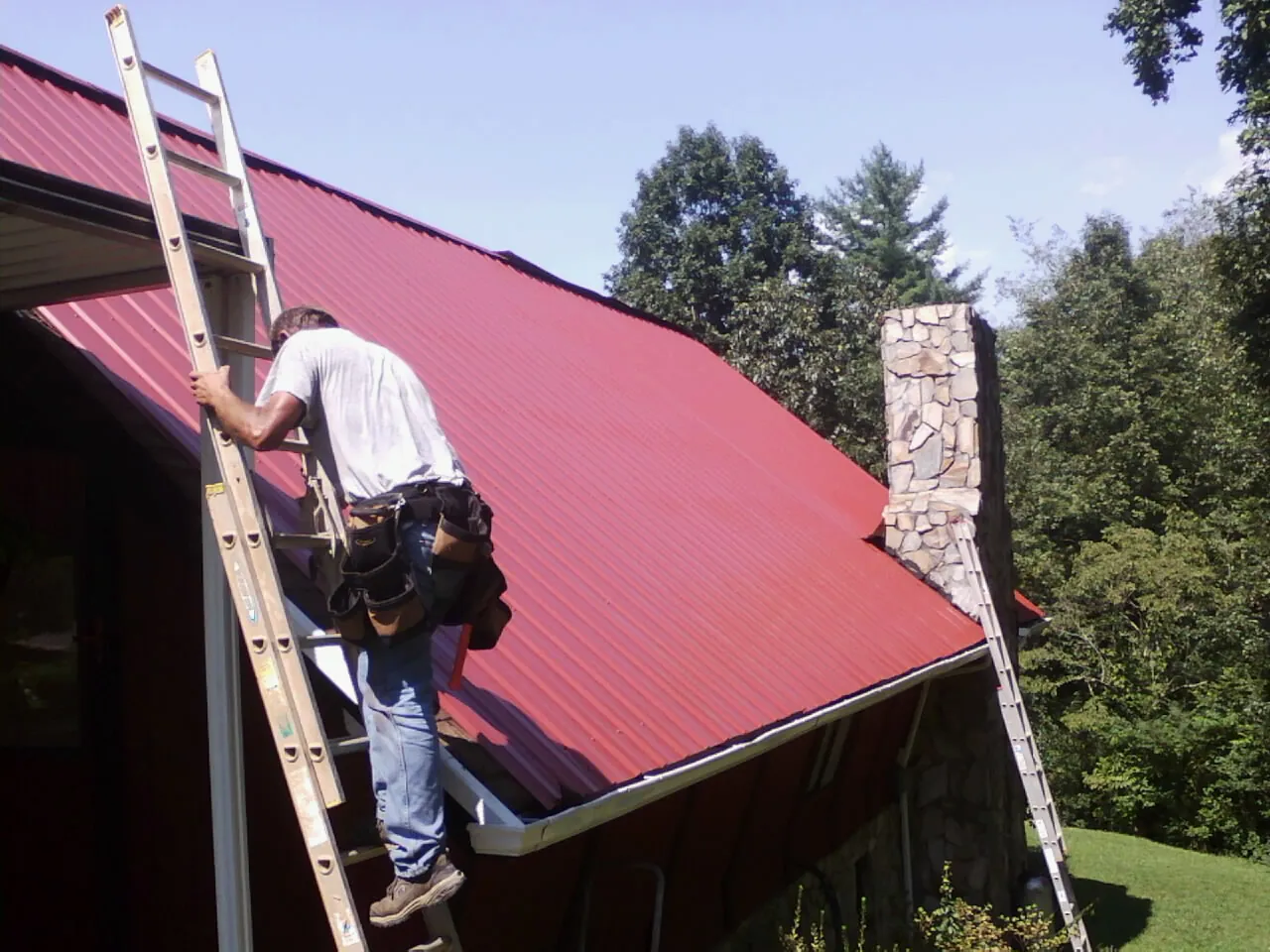 Roofer on a ladder beside a red metal roof and stone chimney — Soffit & Fascia Repair by Roofing Brothers in Sugarcreek